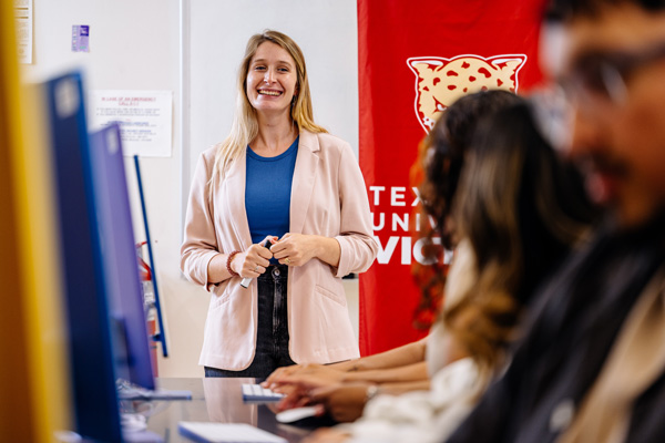 Texas A&M University-Victoria students working at desktop computers during a classroom session, with an instructor standing near a campus-branded banner.