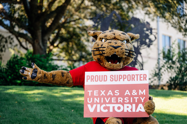 jaX the mascot standing outside holding a Proud Supporter Texas A&M University-Victoria sign