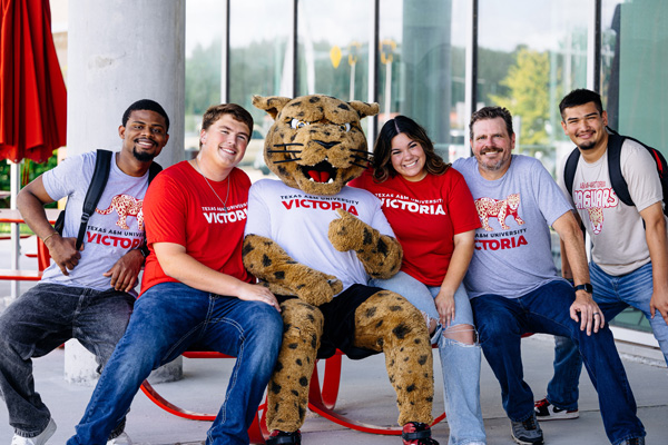 Texas A&M University-Victoria students seated with jaX, the campus mascot, wearing branded T-shirts in front of a glass-windowed building.