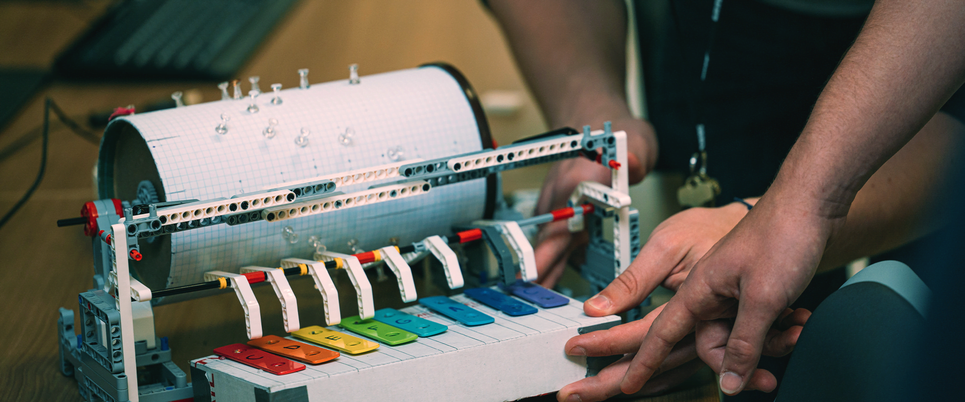 A person adjusts a mechanical device with colorful keys and a cylindrical drum covered in graph paper and pushpins. The machine plays music when the drum rotates