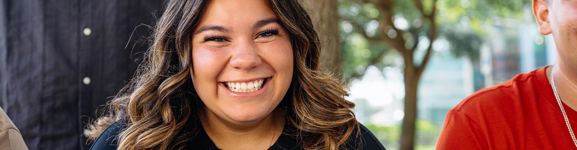Person with long, wavy brown hair smiling broadly and wearing a black shirt. Trees and greenery appear in the background.