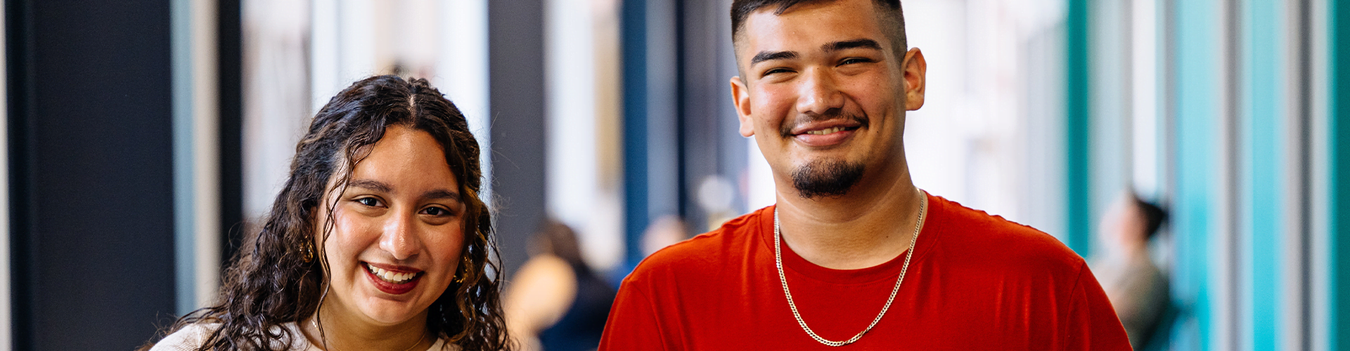 Two people standing indoors in a brightly lit hallway, facing the camera.