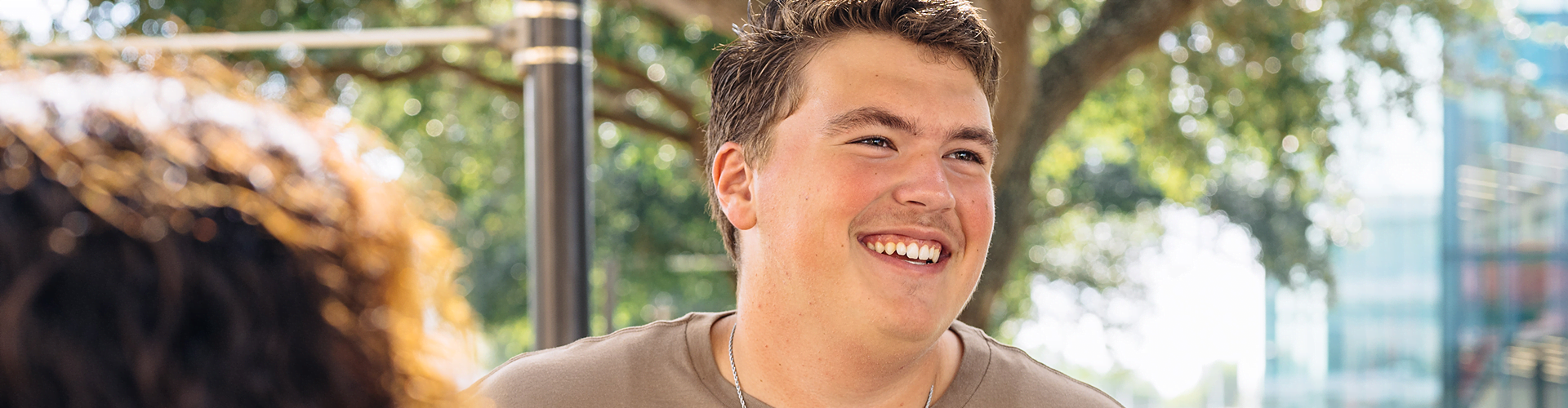 Person smiling outdoors, wearing a light brown shirt and a necklace.