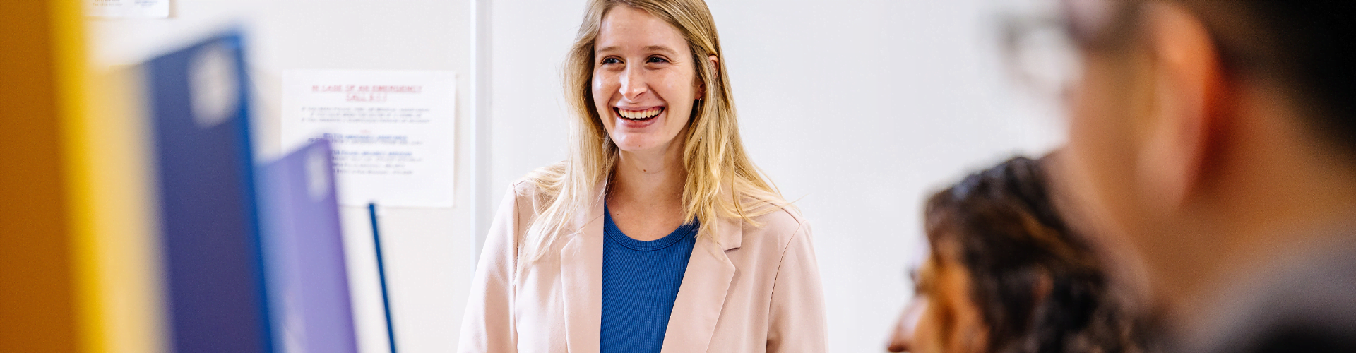 Person standing indoors, smiling and wearing a light pink blazer over a blue top.