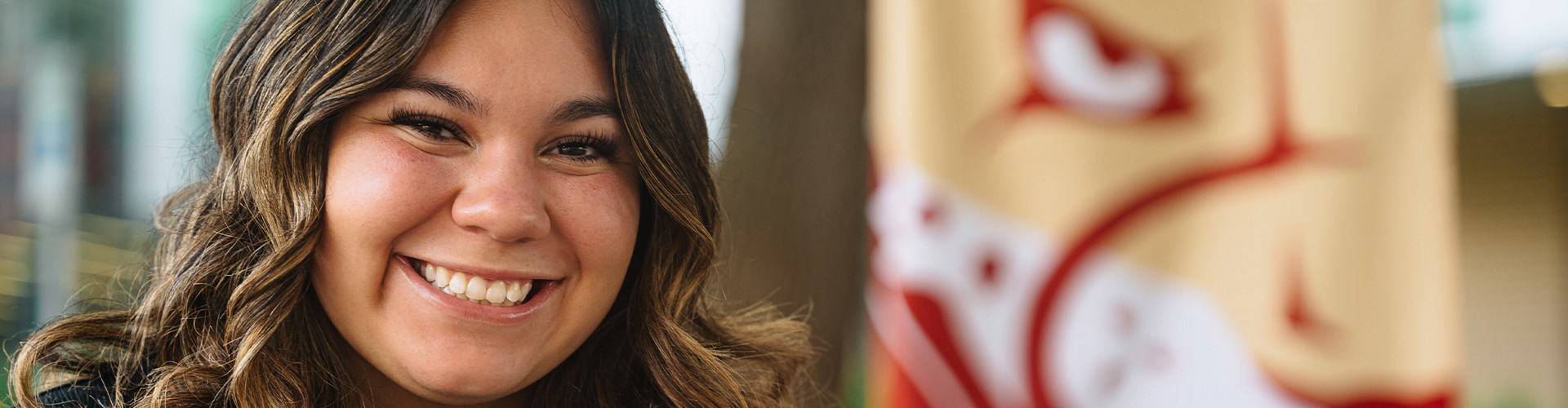 Person with long wavy brown hair smiling, with a blurred flag in the background.