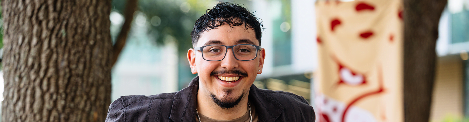 Person with short curly dark hair and glasses, smiling outdoors.