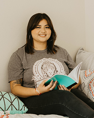 A student sits on a bed, wearing a gray UHV t-shirt and holding an open turquoise-covered book, surrounded by patterned pillows.