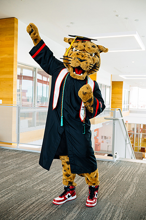 Jax, the Texas A&M University-Victoria jaguar mascot, wearing a graduation gown and cap, with one arm raised in celebration.