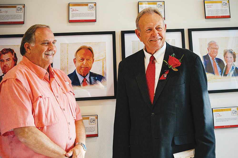 Two men stand in front of a portrait of one of the men.